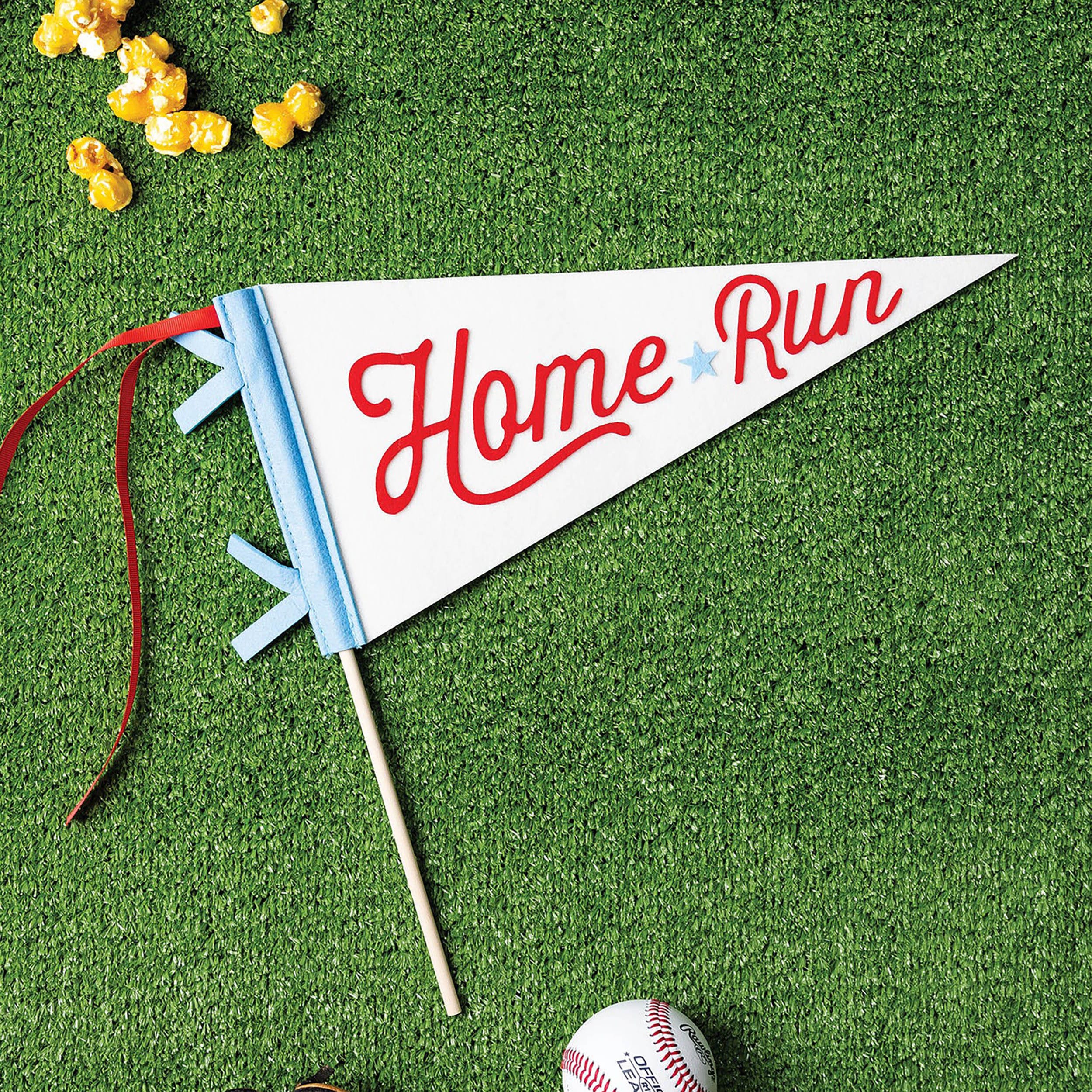 Baseball Pennant Banner - Says Home Run in Red Script - Lifestyle Image of Product on a green turf field - complemented with baseballs, mitt, and caramel corn